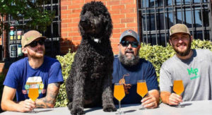 black labradoodle dog sitting with three men drinking beer outside triple c brewing in south end charlotte, n.c.